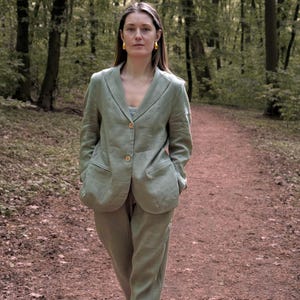 K&ouml;nnte beinhalten: Ein hellgr&uuml;ner Leinenanzug, bestehend aus einem Blazer und einer passenden Hose, wird mit gelben Schuhen getragen. Der Blazer hat zwei Vordertaschen und Holzkn&ouml;pfe. Die Person geht auf einem Weg in einem Wald.