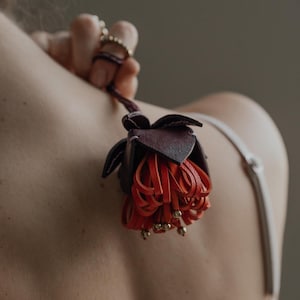 May include: A handcrafted leather flower in shades of burgundy and orange, with gold-colored bead accents. The flower is held by a dark purple leather cord. The image is a close-up shot, highlighting the intricate details of the floral design.