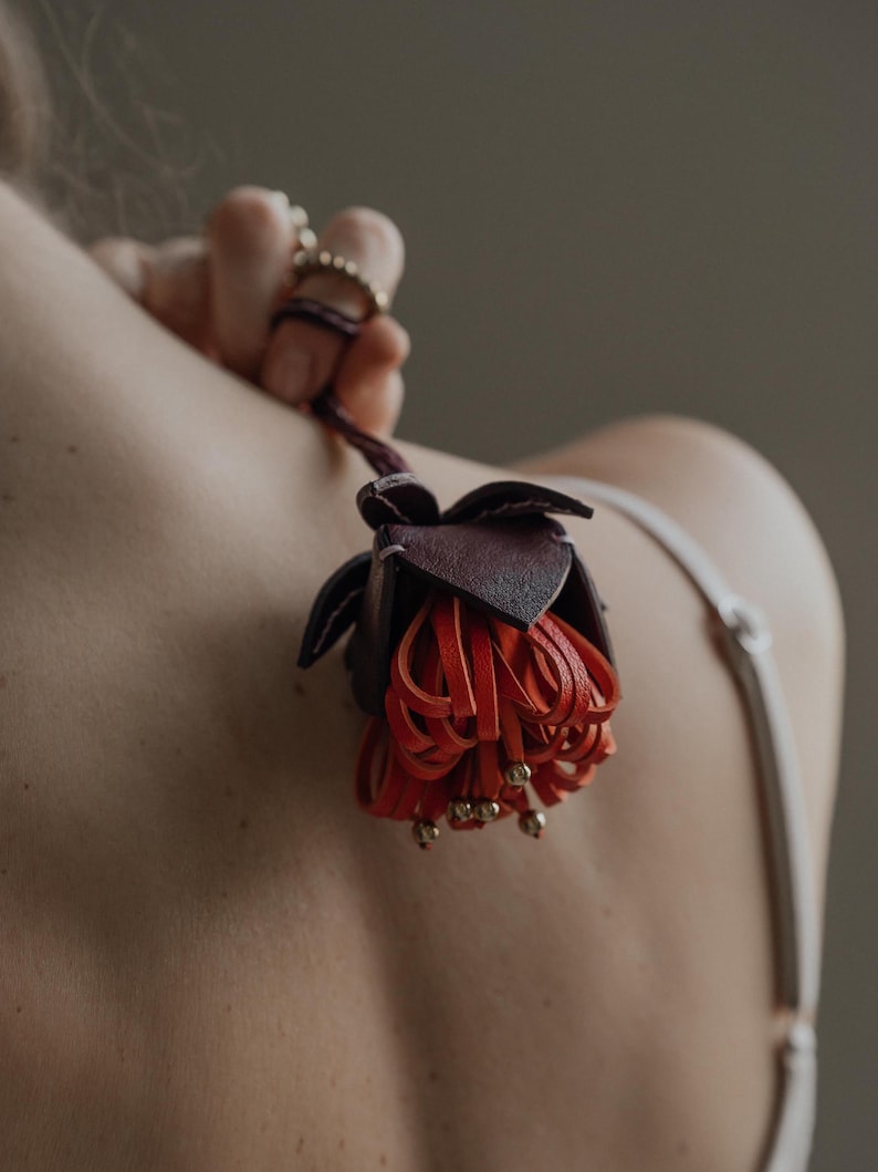 May include: A handcrafted leather flower in shades of burgundy and orange, with gold-colored bead accents. The flower is held by a dark purple leather cord. The image is a close-up shot, highlighting the intricate details of the floral design.