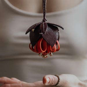 May include: A handcrafted leather flower pendant in shades of burgundy and red, suspended from a thin cord. The pendant features detailed petal and leaf designs, with small gold-colored accents. The overall aesthetic is elegant and artistic.