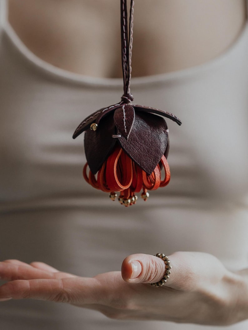 May include: A handcrafted leather flower pendant in shades of burgundy and red, suspended from a thin cord. The pendant features detailed petal and leaf designs, with small gold-colored accents. The overall aesthetic is elegant and artistic.