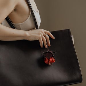 May include: A large, dark brown leather tote bag with a matching strap. A small, decorative leather flower charm in shades of red and brown hangs from the bag. The bag is held by a person wearing a ring.