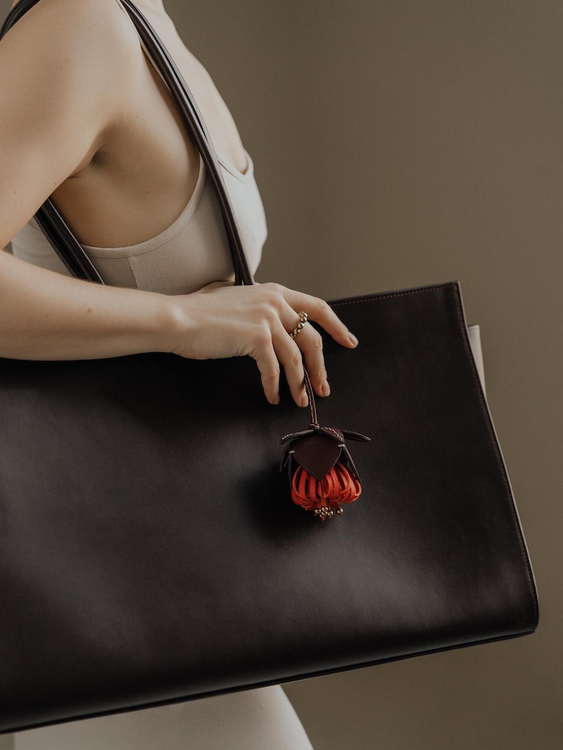 May include: A large, dark brown leather tote bag with a matching strap. A small, decorative leather flower charm in shades of red and brown hangs from the bag. The bag is held by a person wearing a ring.