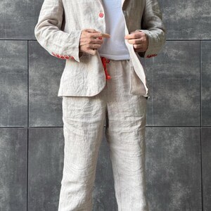 May include: A beige linen suit, consisting of a blazer and pants, with red stitching and buttons. The blazer is open, revealing a white t-shirt underneath. The pants have a drawstring. The suit is made of a lightweight fabric.