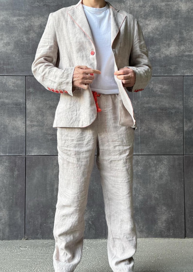 May include: A beige linen suit, consisting of a blazer and pants, with red stitching and buttons. The blazer is open, revealing a white t-shirt underneath. The pants have a drawstring. The suit is made of a lightweight fabric.