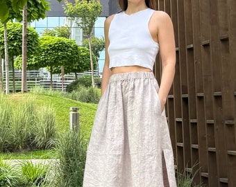 May include: A white crop top and a long, light-coloured linen skirt with a side slit. The skirt has an elastic waistband and pockets. The outfit is worn outdoors on a sunny day.