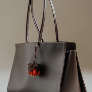 May include: A dark brown leather tote bag with long handles. A decorative flower charm with brown and orange petals hangs from the handle. The bag has a rectangular shape and a minimalist design.