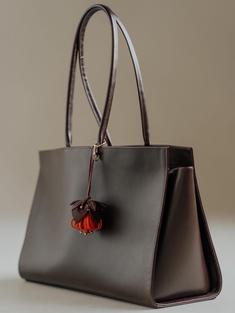 May include: A dark brown leather tote bag with long handles. A decorative flower charm with brown and orange petals hangs from the handle. The bag has a rectangular shape and a minimalist design.