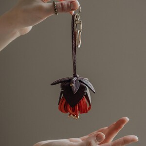 May include: A handcrafted leather keychain in the shape of a fuchsia flower. The keychain features a dark brown top with red fringe petals and gold-colored accents. The keychain is attached to a dark brown leather strap and gold-colored hardware.