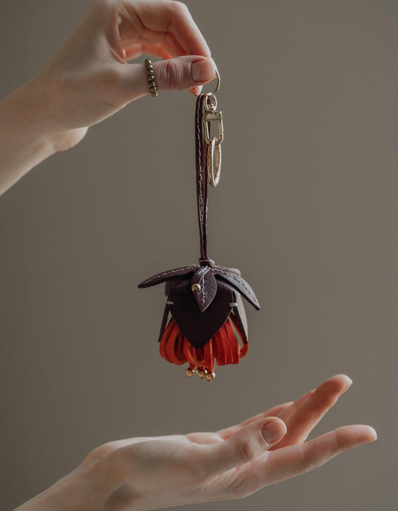 May include: A handcrafted leather keychain in the shape of a fuchsia flower. The keychain features a dark brown top with red fringe petals and gold-colored accents. The keychain is attached to a dark brown leather strap and gold-colored hardware.
