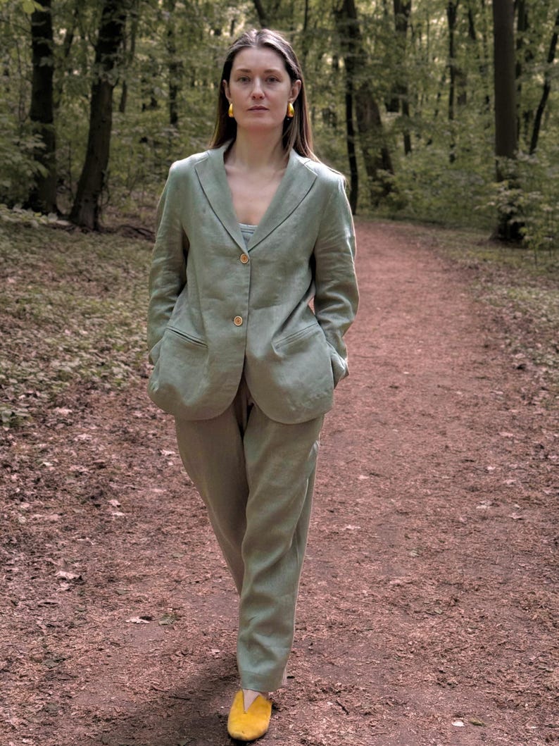 K&ouml;nnte beinhalten: Ein hellgr&uuml;ner Leinenanzug, bestehend aus einem Blazer und einer passenden Hose, wird mit gelben Schuhen getragen. Der Blazer hat zwei Vordertaschen und Holzkn&ouml;pfe. Die Person geht auf einem Weg in einem Wald.