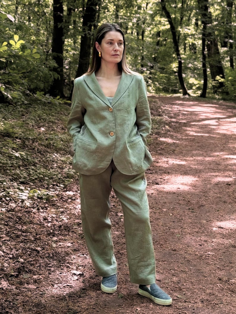 K&ouml;nnte beinhalten: Ein hellgr&uuml;ner Leinenanzug, bestehend aus einem Blazer mit zwei Kn&ouml;pfen und einer passenden Hose. Der Anzug wird mit einem hellblauen Oberteil und dunkelblauen Slip-On-Schuhen getragen. Der Hintergrund ist ein Waldweg.