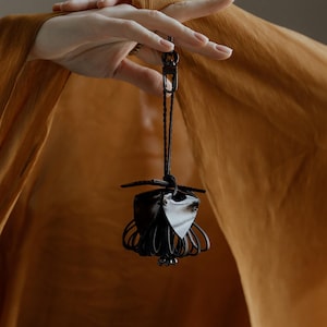 May include: A black leather and metal pendant with a unique, abstract design. The pendant is suspended from a black cord and features a cage-like structure with a central, leaf-shaped element. The background is a warm, golden-brown fabric.