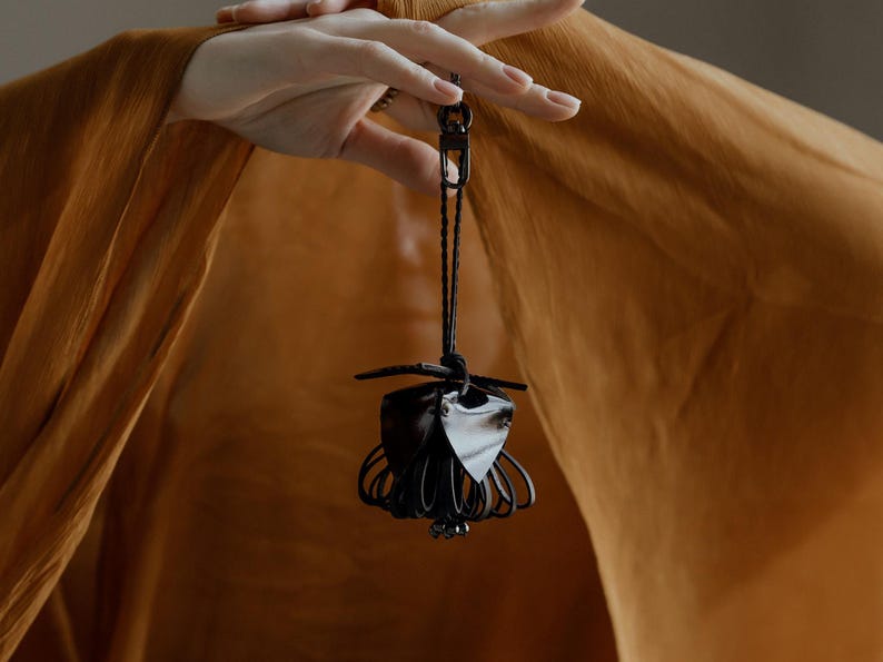 May include: A black leather and metal pendant with a unique, abstract design. The pendant is suspended from a black cord and features a cage-like structure with a central, leaf-shaped element. The background is a warm, golden-brown fabric.