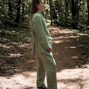 K&ouml;nnte beinhalten: Hellgr&uuml;ner Leinenblazer und passendes Hosen-Set. Der Blazer hat drei Kn&ouml;pfe und die Hose ist gerade geschnitten. Das Outfit wird mit blauen Slip-on-Schuhen getragen. Die Person steht auf einem Waldweg.