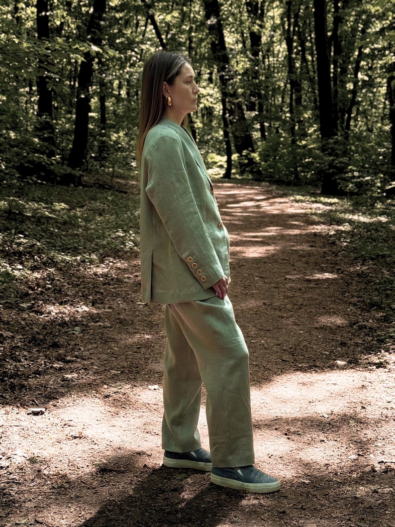 K&ouml;nnte beinhalten: Hellgr&uuml;ner Leinenblazer und passendes Hosen-Set. Der Blazer hat drei Kn&ouml;pfe und die Hose ist gerade geschnitten. Das Outfit wird mit blauen Slip-on-Schuhen getragen. Die Person steht auf einem Waldweg.
