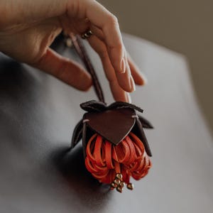May include: A handcrafted leather flower in shades of brown and orange. The flower has a detailed design with multiple petals and small gold-colored beads. The flower is suspended against a dark background.