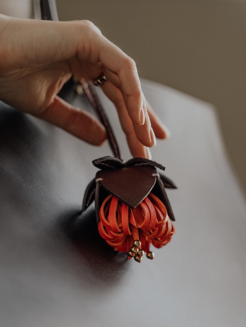 May include: A handcrafted leather flower in shades of brown and orange. The flower has a detailed design with multiple petals and small gold-colored beads. The flower is suspended against a dark background.