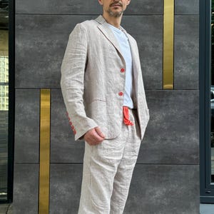May include: A light beige linen suit, consisting of a blazer and matching pants. The blazer has red buttons and a pocket. The person is wearing a white t-shirt underneath. The suit is made of a lightweight fabric, suitable for warmer weather.