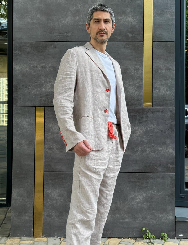 May include: A light beige linen suit, consisting of a blazer and matching pants. The blazer has red buttons and a pocket. The person is wearing a white t-shirt underneath. The suit is made of a lightweight fabric, suitable for warmer weather.