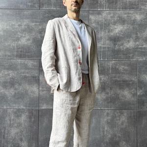 May include: A light beige linen suit, consisting of a blazer and matching trousers, worn over a white t-shirt. The blazer has red stitching and buttons. The outfit is suitable for a casual or semi-formal occasion.