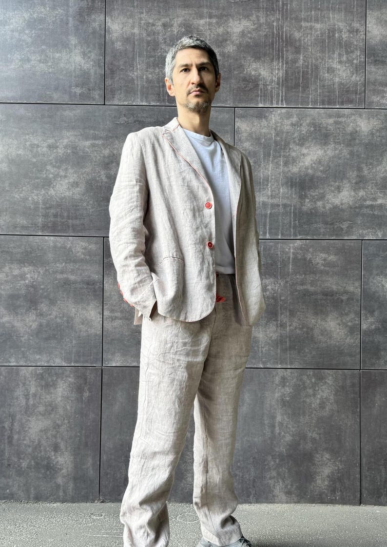 May include: A light beige linen suit, consisting of a blazer and matching trousers, worn over a white t-shirt. The blazer has red stitching and buttons. The outfit is suitable for a casual or semi-formal occasion.