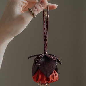May include: A handcrafted leather flower-shaped ornament in shades of burgundy and orange, suspended by a burgundy strap and gold-tone clasp. The flower has layered petals and is accented with small gold beads. The item is held by a hand.