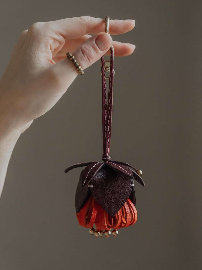 May include: A handcrafted leather flower-shaped ornament in shades of burgundy and orange, suspended by a burgundy strap and gold-tone clasp. The flower has layered petals and is accented with small gold beads. The item is held by a hand.