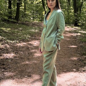K&ouml;nnte beinhalten: Eine Frau in einem hellgr&uuml;nen Leinenanzug, bestehend aus Blazer und passender Hose. Der Anzug hat eine lockere Passform und wird mit dunkelgrauen Slip-On-Schuhen getragen. Der Hintergrund ist eine verschwommene Waldszene mit Sonnenlicht.
