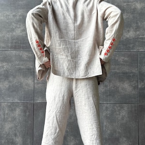 May include: A light beige linen suit, consisting of a blazer and matching pants. The blazer features red buttons on the sleeves. The outfit is made of a textured fabric and is shown from the back.
