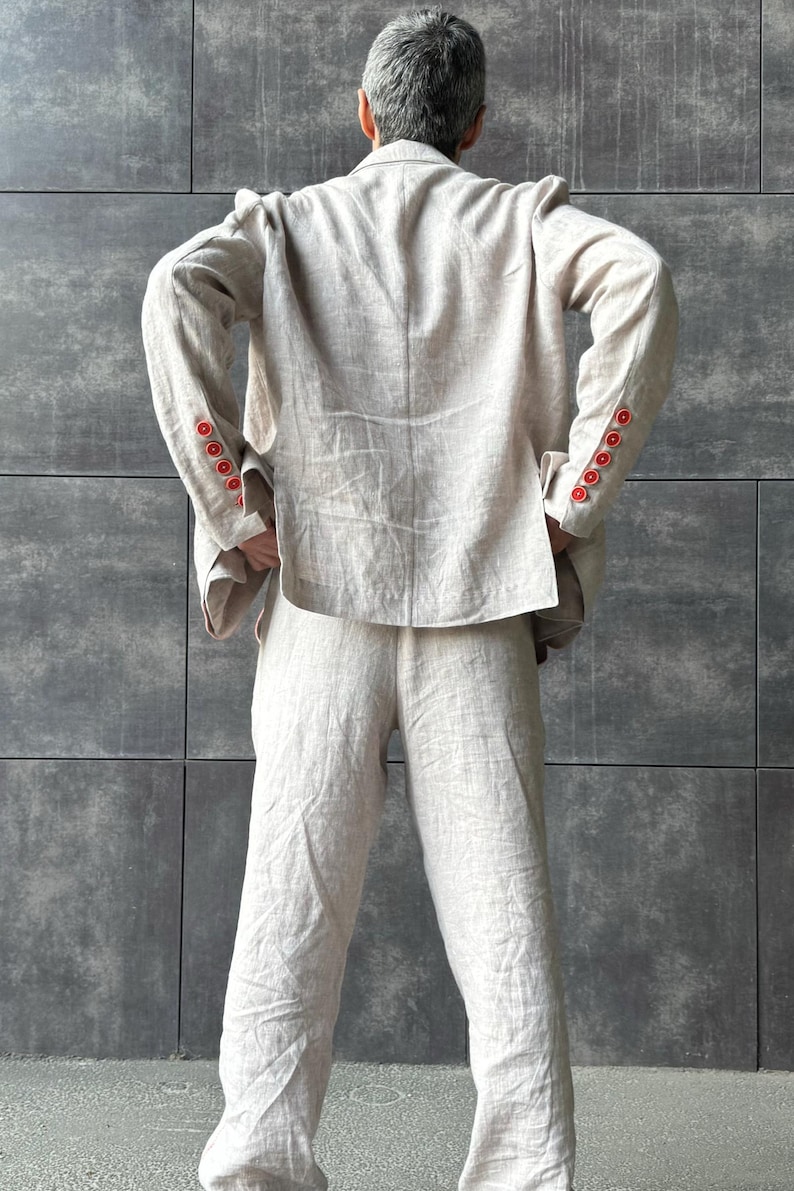 May include: A light beige linen suit, consisting of a blazer and matching pants. The blazer features red buttons on the sleeves. The outfit is made of a textured fabric and is shown from the back.