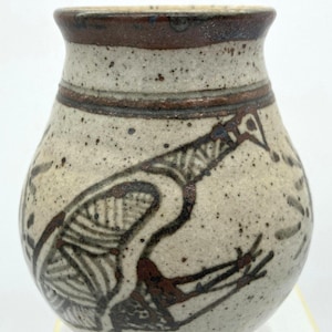 May include: A small, speckled ceramic vase with a brown rim and a decorative bird design. The bird is depicted in brown and white, with intricate line work. The vase has a rounded body and a narrow neck.