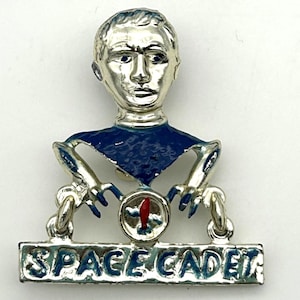May include: Silver-toned Space Cadet pin with a stylized astronaut figure. The pin features a blue collar, blue accents, and the words "SPACE CADET" in blue lettering. A red and white emblem is centered on the pin.
