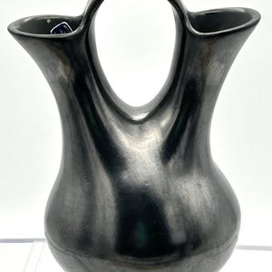 May include: A black pottery vase with a unique double-spout design and a large handle. The vase has a smooth, polished surface and a rounded base. The dark color and elegant shape suggest a handcrafted item, suitable for home decor.