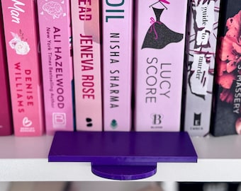May include: A purple book stand with a rectangular top and a semi-circular base, positioned in front of a row of books. The books have pink covers with titles like "Lucy Score" and "Ali Hazelwood".