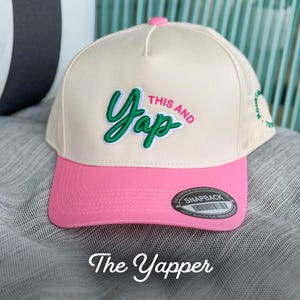 Peut inclure: Une casquette de baseball beige et rose avec un logo brodé vert et blanc qui dit "This and Yap". La casquette a une face inférieure rose et une étiquette noire qui dit "Snapback" à l'arrière.