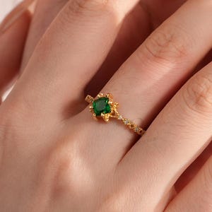 Könnte beinhalten: Ein goldener Ring mit einem zentralen smaragdgrünen Edelstein, der in einem floralen Design gefasst ist. Der Ring befindet sich an einem Finger, wobei die Hand im Fokus steht. Das Band des Rings hat ein zartes, strukturiertes Muster. Der Hintergrund ist verschwommen, mit einer weichen, neutralen Farbpalette.