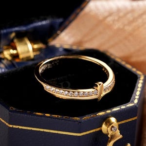 May include: A gold ring with small diamonds set in a band, presented in a navy blue hexagonal jewellery box with gold trim. The ring is the focal point, set against a black interior. The box is open, revealing the ring.