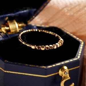 May include: A gold ring with small square beads is displayed inside an open navy blue hexagonal jewellery box. The box has gold trim and a gold clasp. The ring is set against a black velvet interior.