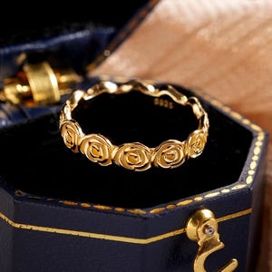 May include: A gold-colored ring with a floral design, presented in a dark blue hexagonal jewelry box with gold trim. The ring features a series of rose-shaped embellishments. The box is open, revealing the ring against a black interior.