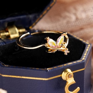 Puede incluir: Un anillo de oro con diseño de mariposa se muestra dentro de una caja de joyería azul. El anillo tiene una delicada mariposa con piedras amarillas y transparentes. La caja abierta revela un interior negro y detalles dorados.