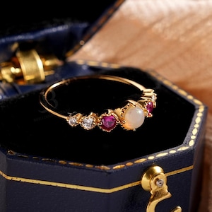 May include: A gold ring featuring an opal centre stone, with red and clear gemstones, sits in an open, dark blue hexagonal jewellery box. The box has a black interior and gold detailing. A peach-coloured fabric forms the background.