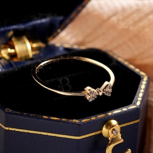 Puede incluir: Un anillo de oro con diseño de lazo, presentado en una caja de joyería hexagonal azul oscuro con ribetes dorados. El anillo presenta pequeñas piedras brillantes. La caja está abierta, revelando el anillo sobre un interior de terciopelo negro.
