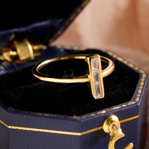 Op de afbeelding: Een gouden ring met een rechthoekige, heldere edelsteen wordt getoond in een open, marineblauwe, zeshoekige sieradendoos. De doos heeft gouden randen en een gouden sluiting. De achtergrond omvat zachte, neutraal getinte stof en kleine witte bloemen.