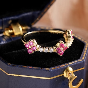 Peut inclure: Une bague en or ornée de pierres roses et de diamants blancs, présentée dans un écrin bleu. La bague présente un motif floral avec trois groupes de fleurs roses. L'écrin est ouvert, révélant la bague sur un fond noir.