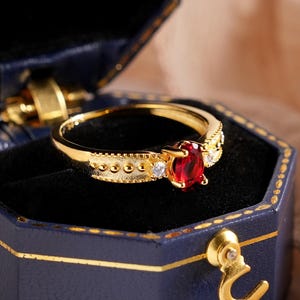 May include: A gold ring with a red gemstone, presented in a blue hexagonal jewellery box. The ring has a central red stone and smaller clear stones. The box is navy blue with gold trim and a gold clasp.