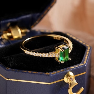 Könnte beinhalten: Ein goldener Ring mit einem rechteckigen grünen Edelstein, präsentiert in einer dunkelblauen sechseckigen Schmuckschatulle. Der Ring ist mit kleinen, klaren Steinen verziert. Die Schatulle hat goldene Verzierungen und einen goldenen Verschluss. Weiße Blumen sind im Vordergrund.