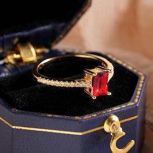 Op de afbeelding: Een gouden ring met een rechthoekige rode edelsteen wordt getoond in een open, marineblauwe, achthoekige sieradendoos. De ring heeft een delicate band met kleine, sprankelende accenten. De doos is gevoerd met zwart fluweel en heeft gouden randen.