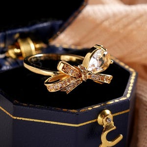 May include: A gold ring with a bow design, featuring a large clear gemstone and smaller accent stones. The ring is displayed in a navy blue hexagonal jewellery box with gold trim. The box is open, revealing a black interior.