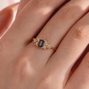 May include: A gold ring with a rectangular, dark blue gemstone and smaller clear stones. The ring has an ornate design with a thin gold band. The ring is a piece of jewellery. The ring is a piece of jewellery.